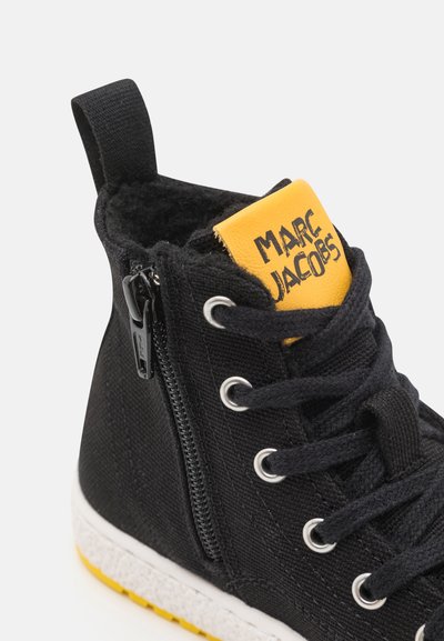 Sneaker alta nera in materiale di tela, con etichetta gialla del marchio, occhielli in argento, chiusura con zip e suola bianca testurizzata con accenti gialli.