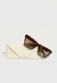 Barna acetát napszemüveg geometrikus macskaszem formával, átmenetes lencsékkel, krémszínű, háromszög alakú, dombornyomott logóval ellátott tokon pihenve.