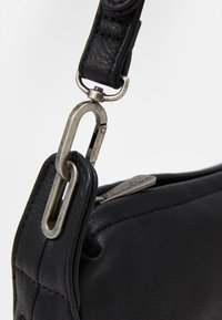 Černý kožený popruh na tašce se stříbrnou kovovou sponou. Vyznačuje se texturovaným povrchem a detaily šití. Detailní pohled na kovové prvky a materiál.