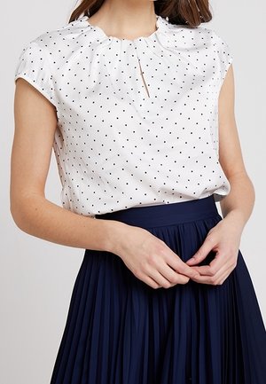 Frau trägt eine weiße kurzärmelige Bluse mit Polka-Dots, einem gerafften Ausschnitt und einem marineblauen Plissee-Rock.