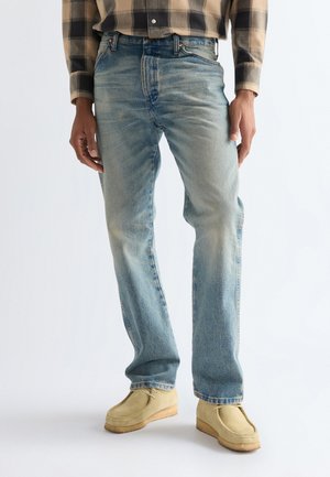 Person, die ausgewaschene blaue Jeans mit geradem Bein, beige Wildleder-Mokassins und ein braunes kariertes Hemd trägt, steht vor einem weißen Hintergrund.