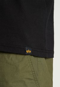 Svart bomull T-skjorte med en synlig hemlabel som har en geometrisk logo i oransje, sammen med olivengrønne bukser som viser en sidesøm.