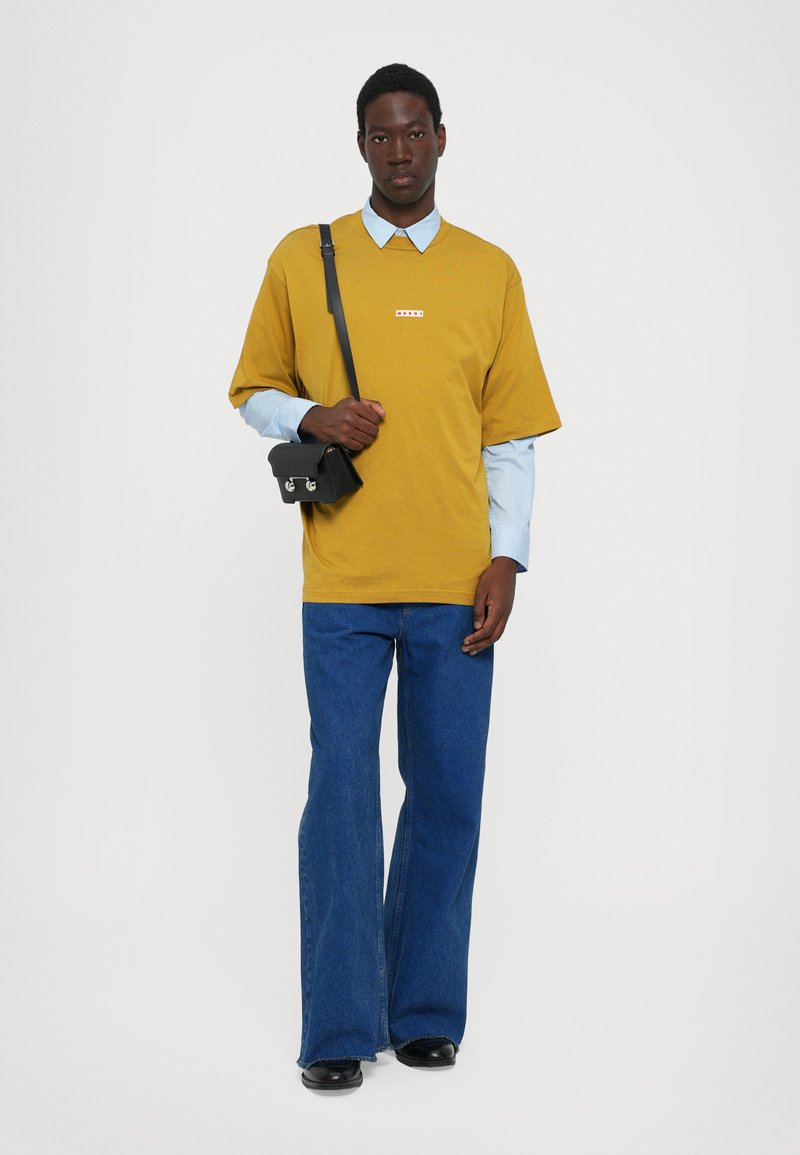 Mustard yellow oversized t-shirt with a small red logo, layered over a light blue collared shirt, paired with flared blue jeans and a black bag.