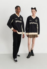 Jerseys de manga larga en negro y crema con cuello en V, que presentan el texto "yourturn". Pantalones negros, falda de cuadros y botas de combate completan los atuendos.