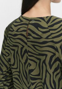 Grön och svart randig topp med zebra-mönster och slits i ryggen, med en metallring för stängning. Mjuk tyg med en slät yta och avslappnad passform.