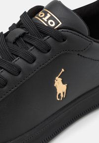 Černá kožená teniska se zlatým logem, strukturovaným svrškem, plochými černými tkaničkami a gumovou podrážkou s malými perforacemi podél okraje.