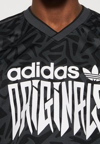Camiseta deportiva negra con un patrón geométrico, diseño de cuello en V, que presenta un destacado texto en blanco "adidas ORIGINALS" y acentos de marca clásicos.