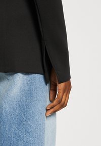 Czarna kurtka z teksturowanym wykończeniem, z bocznymi rozcięciami u dołu, zestawiona z jasnoniebieskimi dżinsami. Zbliżenie na rękę przy ubraniu.