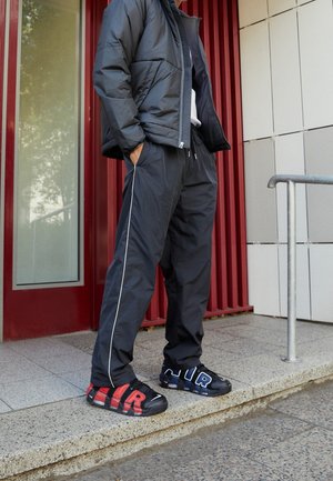 Osoba stojąca z rękami w kieszeniach, ubrana w czarną kurtkę puchową, czarne spodnie dresowe z bocznym paskiem oraz niepasujące do siebie czarne trampki z czerwonym i niebieskim napisem "AIR".