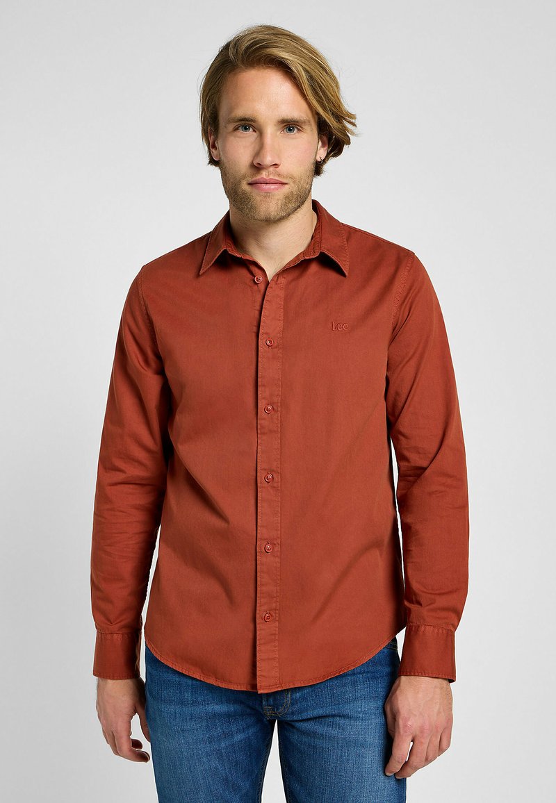 Jonge man met lichtbruin haar, gekleed in een roestkleurig overhemd met knopen en blauwe jeans, staand tegen een effen lichte achtergrond.
