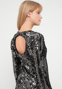 Μαύρο φόρεμα με παγιέτες και μακριά μανίκια, που διαθέτει κοπή στη πλάτη σε στρογγυλό σχήμα, υφασμάτινη υφή και εφαρμοστή σιλουέτα. Οι λαμπερές παγιέτες δημιουργούν μια ανακλαστική επιφάνεια.