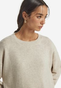 Helles, beiges Strickpullover mit geripptem Rundhalsausschnitt und lockerer Passform. Weiche Textur, die die Schlichtheit des Designs hervorhebt.