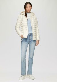Veste rembourrée crème avec capuche, dotée d'une fermeture éclair à l'avant et de poches latérales, portée sur un haut à plusieurs couches bleu clair et un jean clair, avec des chaussures blanches.