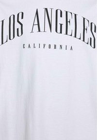 T-shirt blanc avec un texte noir en gras indiquant "LOS ANGELES" en arc au-dessus de "CALIFORNIA", avec différentes tailles et styles de police. Texture de tissu douce.