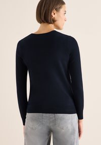 Maglione aderente blu navy con maniche lunghe e scollatura rotonda, realizzato in tessuto morbido, dotato di orlo e polsini a coste. Indossato con jeans grigi.
