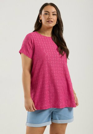 Donna con lunghi capelli scuri che indossa orecchini a cerchio dorati, una maglietta rosa a maniche corte e texture, e pantaloncini di jeans azzurri risvoltati.