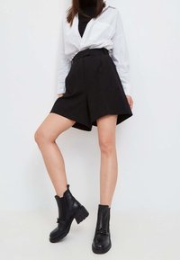 Botas de tobillo de cuero negro con suelas gruesas y herrajes decorativos, combinadas con una camisa blanca de botones y pantalones cortos negros a medida.