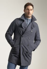 Gabardina gris oscuro de doble botonadura con grandes botones, cuello con muesca y bolsillos laterales, usada sobre un suéter azul claro.