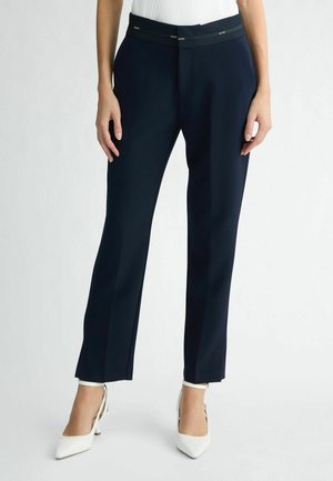 Navy blue tailored pants with a smooth texture, flat front design, and elastic waistband featuring brand logo. Paired with white pointed heels.