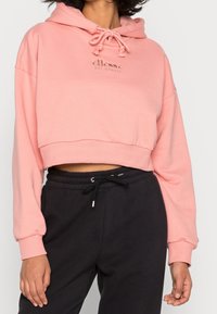 Růžová zkrácená mikina s kapucí na stažení, žebrovanými manžetami a logem na hrudi, spárovaná s černými tepláky na stažení.