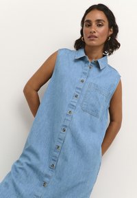 Un vestido de mezclilla sin mangas, de color azul claro, con un cuello tipo camisa, cierre frontal de botones y un único bolsillo en el pecho. Textura sutil visible.