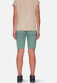 Beige kortærmet skjorte parret med petrolfarvede knælange shorts. Shortsene har to baglommen og folded kant. Sorte fodtøj er synlige.