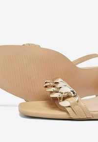 Sandalias de tacón beige con un acabado brillante, que presentan un acento de cadena a lo largo de la correa delantera y una suela de goma texturizada.