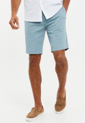 Pantalones cortos de algodón azul claro con frente plano, dobladillos doblados y bolsillos laterales, combinados con zapatos marrones tipo slip-on. Camisa blanca de botones.