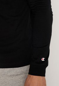 Schwarzes langärmeliges T-Shirt aus Baumwolle mit figurbetontem Schnitt. Auffälliges rotes und weißes Logo auf der Manschette; kombiniert mit grauen Jogginghosen.