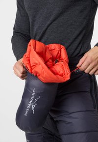 Eine rote isolierte Jacke wird in einen dunkelgrauen Packsack mit einem Logo gepackt, der eine glatte Textur und einen Kordelzugverschluss hat.