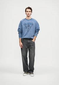 Sudadera gráfica azul con un estampado triangular y el texto "GUESS"; combinada con jeans rectos de color gris oscuro y zapatillas blancas.