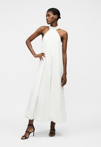 Femme debout, une main sur la hanche, portant une robe maxi blanche à col licou sans manches et des sandales noires à talons hauts.