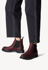 Botines de cuero burdeos con costados elásticos negros, puntera redonda, tacón bajo y suela de goma texturizada. Jeans con bajo recortado visible.