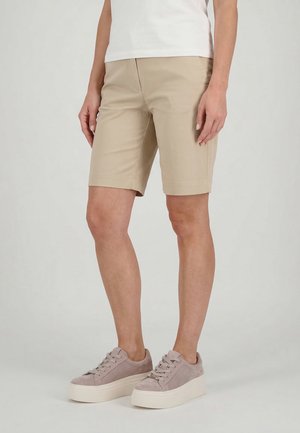 Osoba ubrana w beżowe krótkie spodenki do kolan, biały top i jasnoróżowe trampki na platformie, stojąca na jednolitym tle.