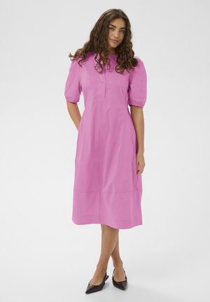 Žena s kučeravými vlasmi v ružových šatách po kolená s nadýchanými rukávmi a čiernymi lodičkami so zadným pásikom, stojaca na bielom pozadí.