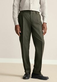 Grüne Tweed-Hose mit geradem Schnitt, mit Falten vorne und Knopfverschluss. Kombiniert mit schwarzen Schuhen und einem hellgrauen Hemd.