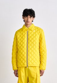 Casaco amarelo acolchoado com gola alta, fecho de correr completo e padrão de losangos. Usado com calças amarelas a condizer, exibindo um visual coeso.