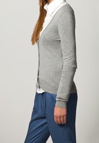 Světle šedý cardigan s designem do V, žebrovanými manžetami, předními knoflíky a měkkou texturou, nošený přes bílou košilovou košili a modré kalhoty.