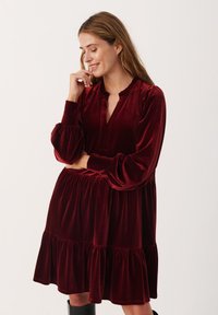 Abito in velluto borgogna con scollo arricciato, maniche lunghe a sbuffo e design a gonna a strati. Presenta una linea dritta e una superficie liscia.