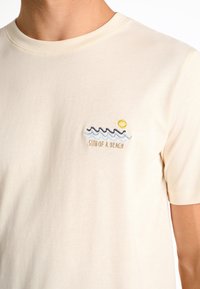 Beige-cotton t-paita, jossa on pieni brodeerattu kuvio aalloista ja auringosta, ja sen alla teksti "SUN OF A BEACH".