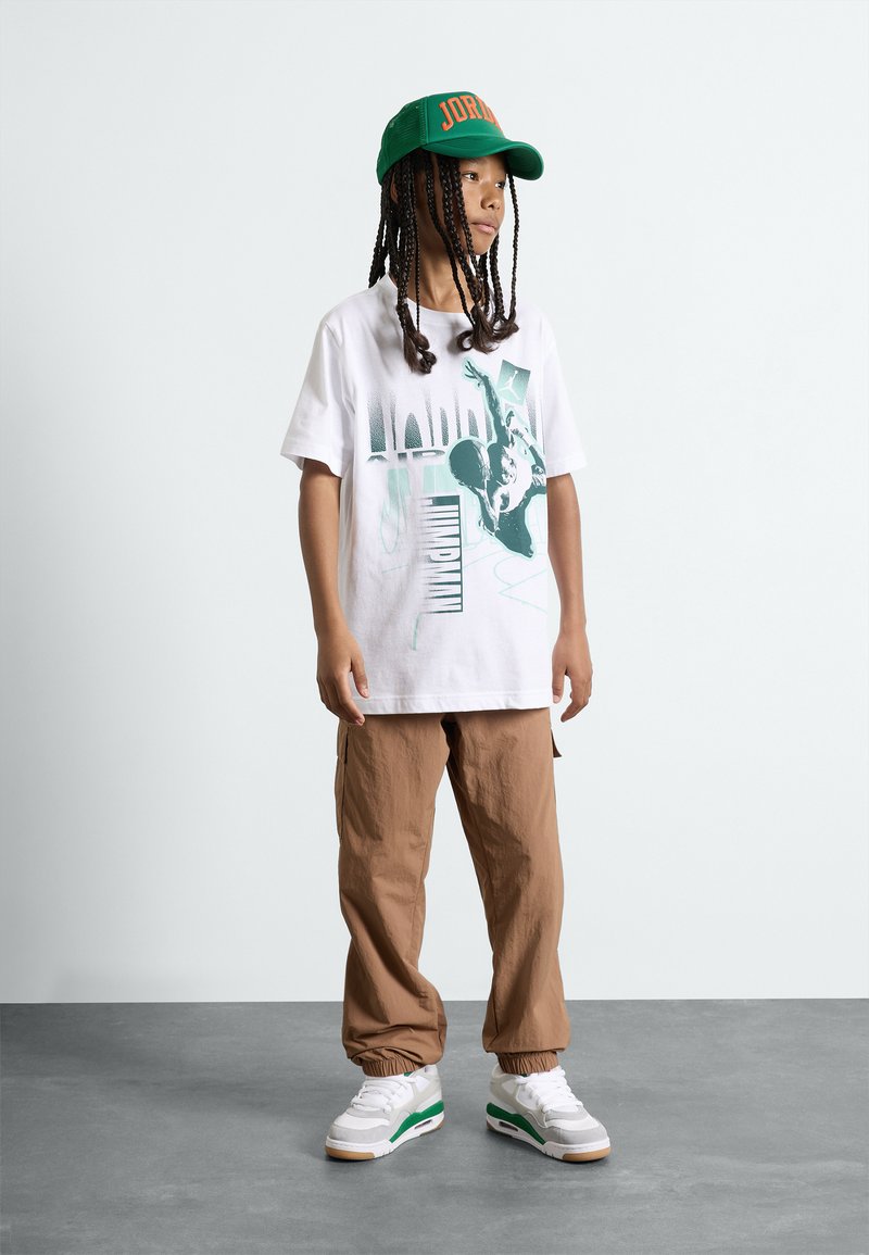 Camiseta gráfica blanca con estampado verde, pantalones cargo beige con puños elásticos y zapatillas blancas con detalles verdes. Gorra verde con el texto "JORDAN".