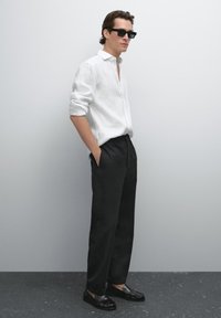 Man wearing black sunglasses, white long-sleeve button-up shirt with rolled sleeves, black trousers, and black loafers standing against gray wall.