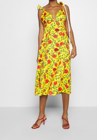 Vestido midi amarillo con estampado floral, con escote en V y tirantes con volantes, que presenta patrones de rosas rojas y hojas verdes, combinado con tacones rojos con tiras.