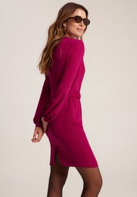 Fuksjowa sukienka z długimi bufkami, przewężona w talii, z wycięciem po boku. Gładki materiał i nowoczesny krój uzupełniają duże czarne okulary przeciwsłoneczne.
