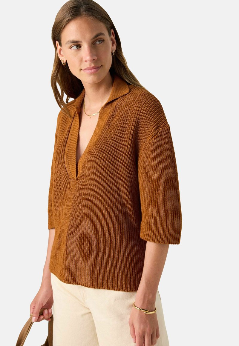 Femme portant un pull en tricot couleur rouille avec des manches 3/4, un pantalon crème, des bijoux en or et tenant un sac fourre-tout marron, regardant sur le côté.