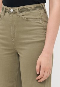 Kvinne høyre hånd hviler ved siden av kaki bukser med strukturert midjebånd detaljer og metall knapp lukking.