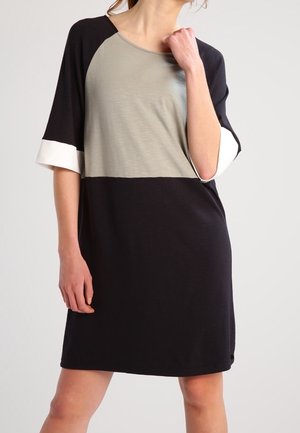 Kobieta ubrana w sukienkę do kolan z czarnym dołem, beżowym topem i czarnymi rękawami z białymi mankietami, stojąca na jednolitym tle.