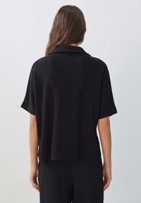 Černý krátký rukáv s límečkem a volným střihem, vyrobený z texturované látky, s mírně zakřiveným lemem a spadlými rameny.
