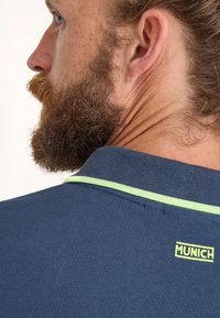 Marinblå pikétröja i en strukturerad tygkvalitet, med en grön detalj längs kragen och en liten gul "MUNICH"-logotyp på baksidan.