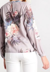 Bluse med blomstermønster i blødt lavendelfarvet stof med rosa, hvide og grønne blomster, lange ærmer og detajle med knapper på nakken bagtil. Stylet med hvide bukser.